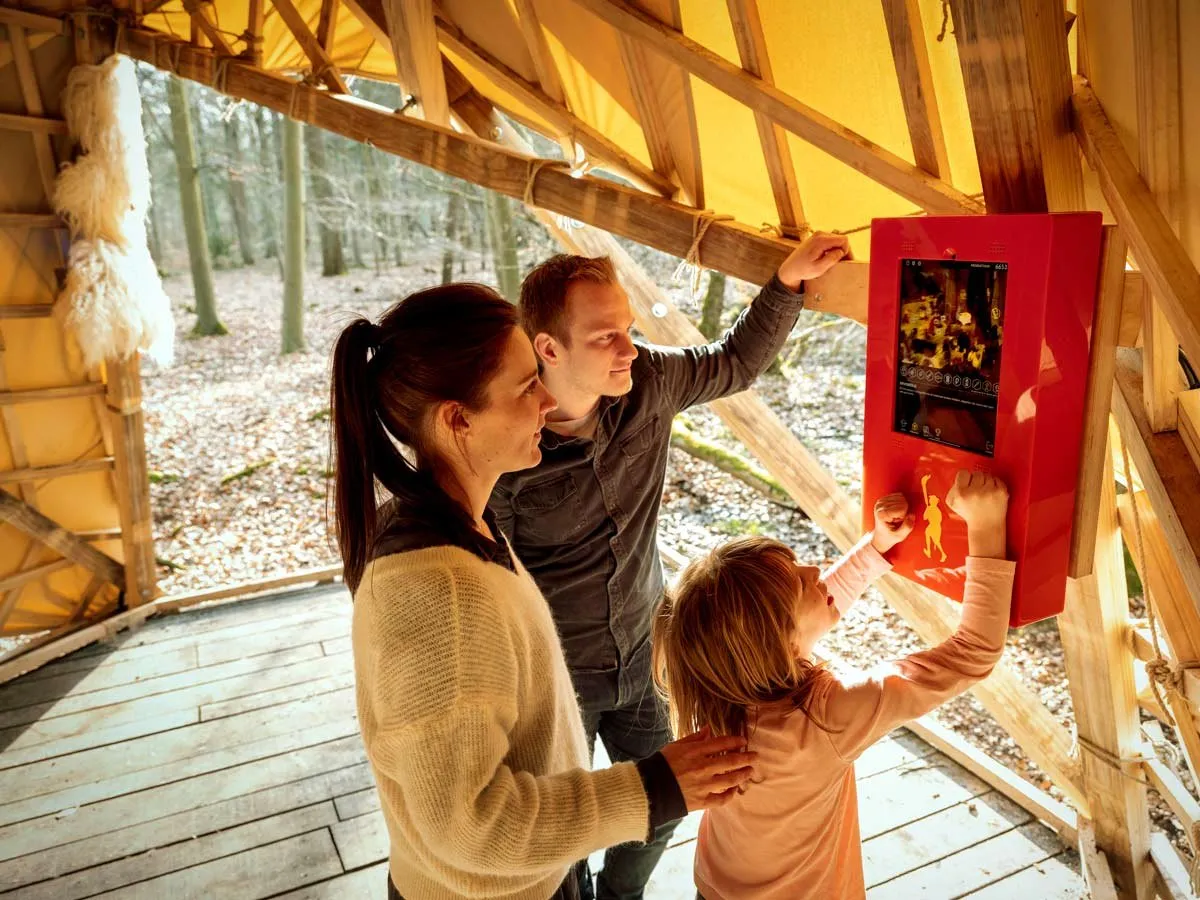 Een gezin speelt samen op een rood interactief station in een houten hut in het bos.
