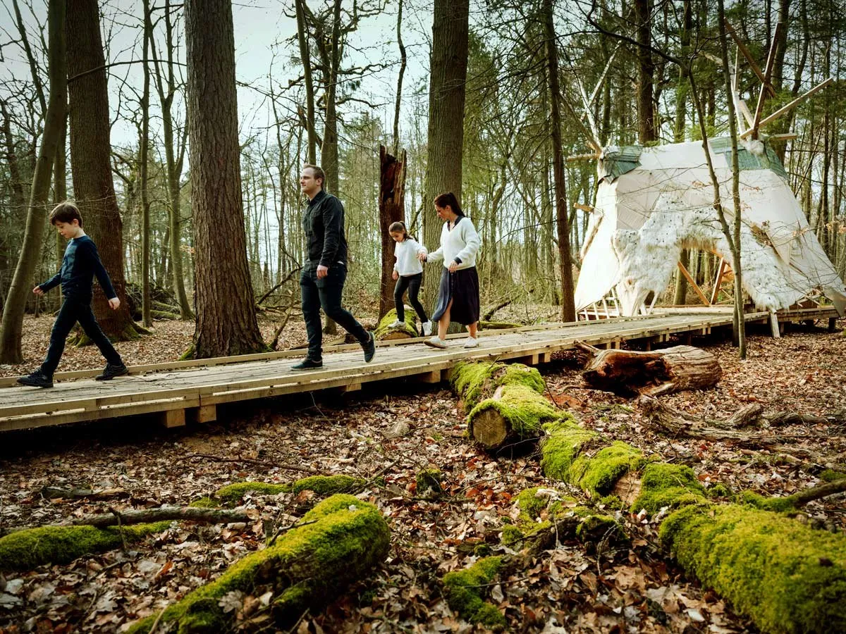 Bezoekers lopen over een houten vlonderpad door het bos naar een tentconstructie.