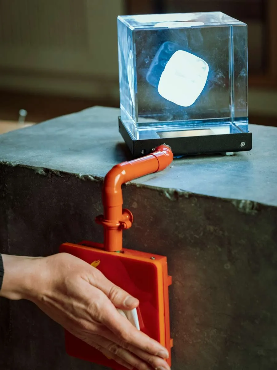 Een hand bedient een rood apparaat onder een verlichte glazen kubus.
