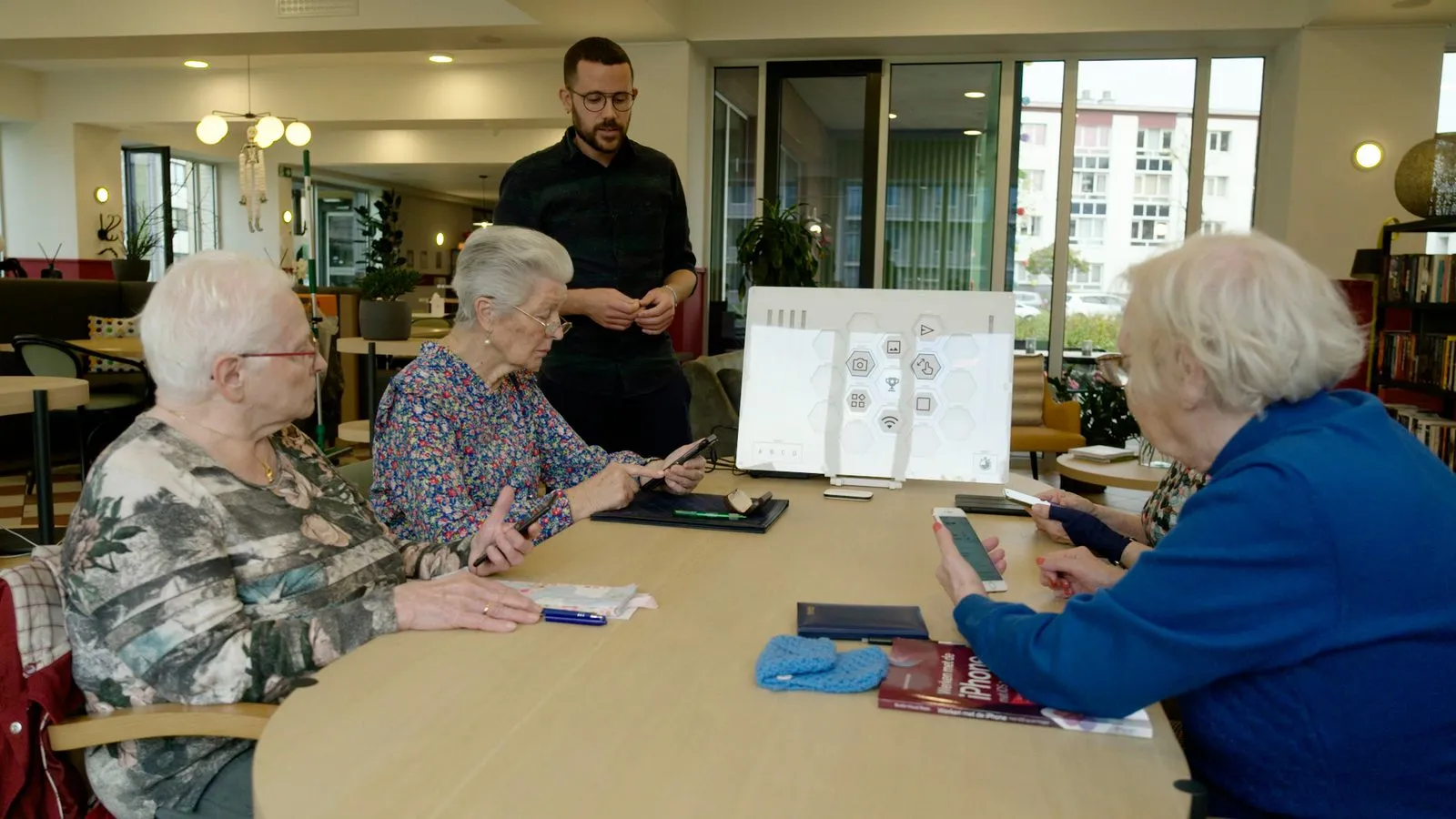 Een lesgever staat bij drie oudere cursisten met smartphones; op tafel staat het Hiveboard met oplichtende zeshoeken.
