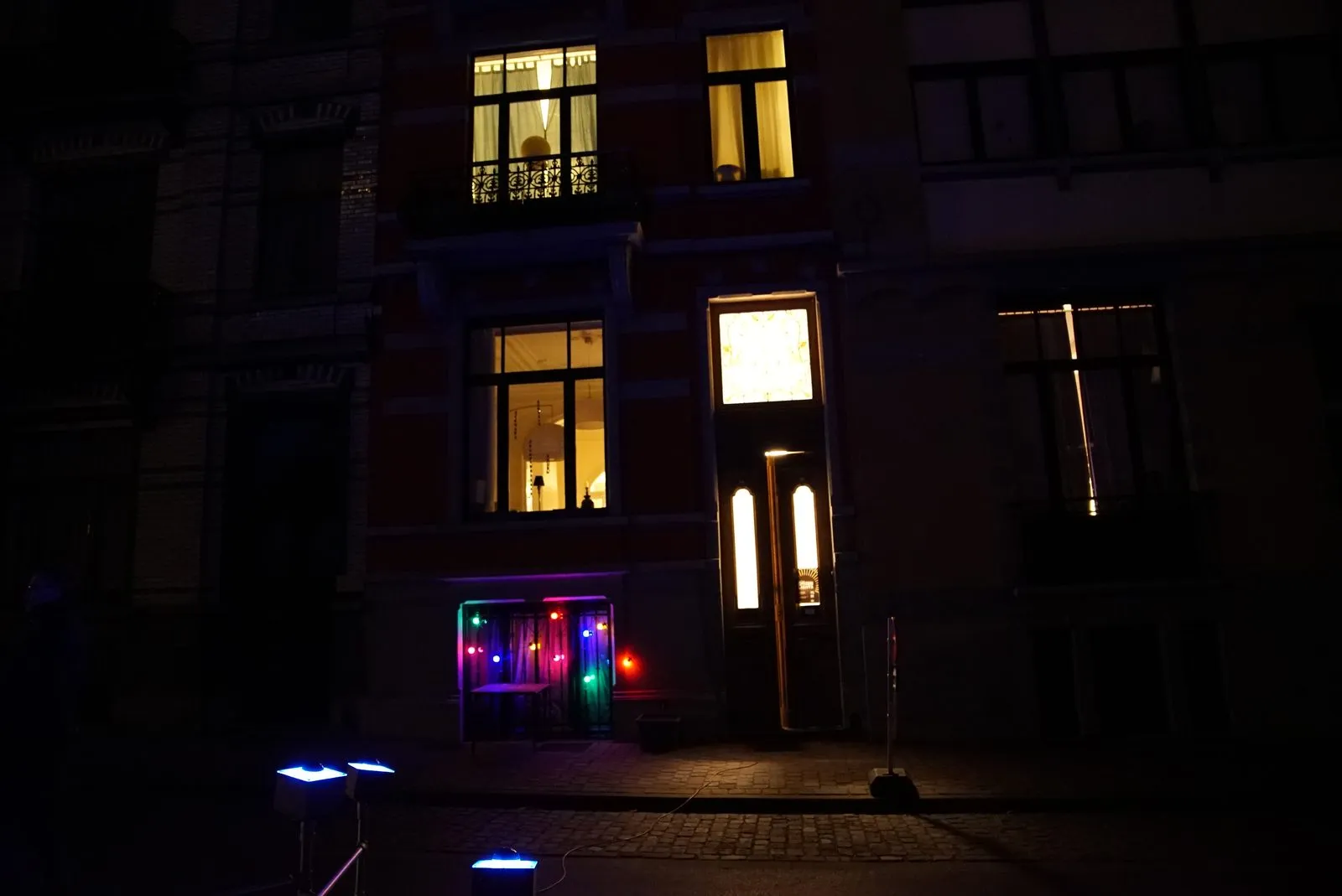 De straat bij nacht: achter een raam op de begane grond branden gekleurde lampjes, op straat staan blauw oplichtende knoppen.