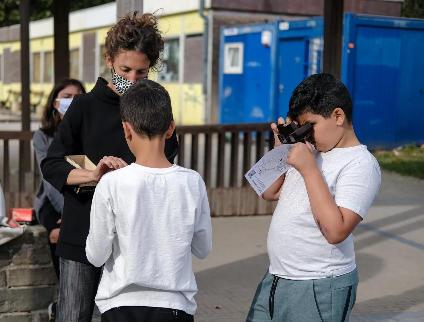 Twee kinderen kijken door een kijkkastje op statief, een begeleidster kijkt mee, aan de rand van een bouwterrein.