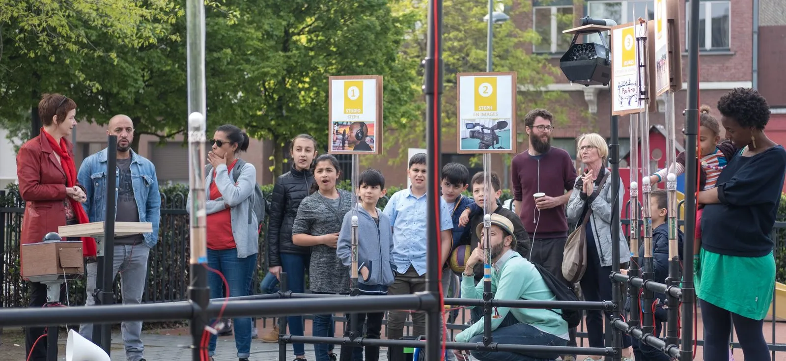 Op een plein staat een installatie van zwarte buizen met genummerde bordjes; kinderen en volwassenen kijken toe.