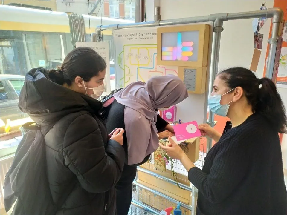 Drie vrouwen bij een mobiele stemunit op een buizenframe, met een gekleurd ledscherm en een roze stemkaart.