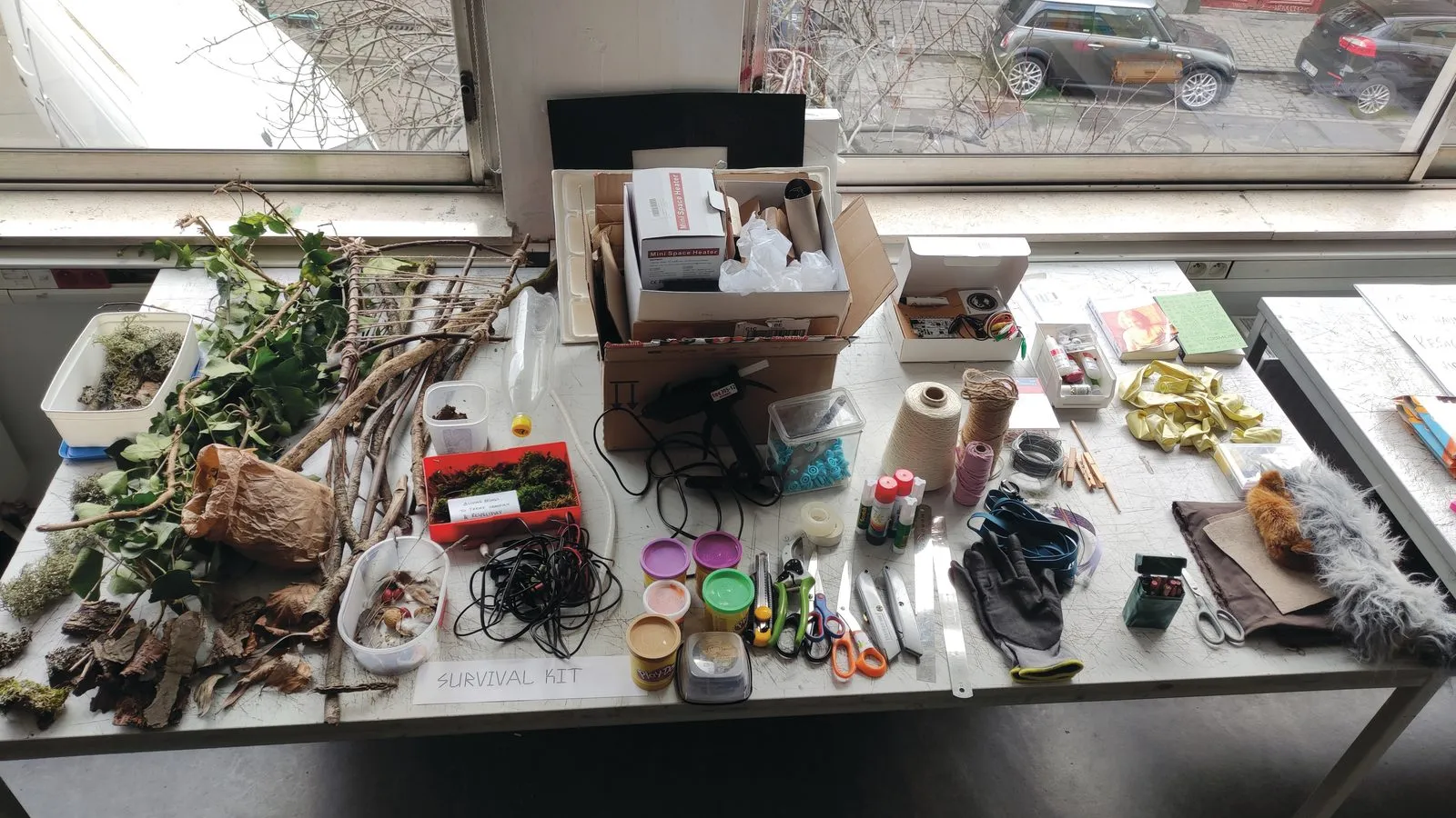 Een werktafel vol materiaal, van takken en mos tot lijmpistool en garen, met een bordje “survival kit”.