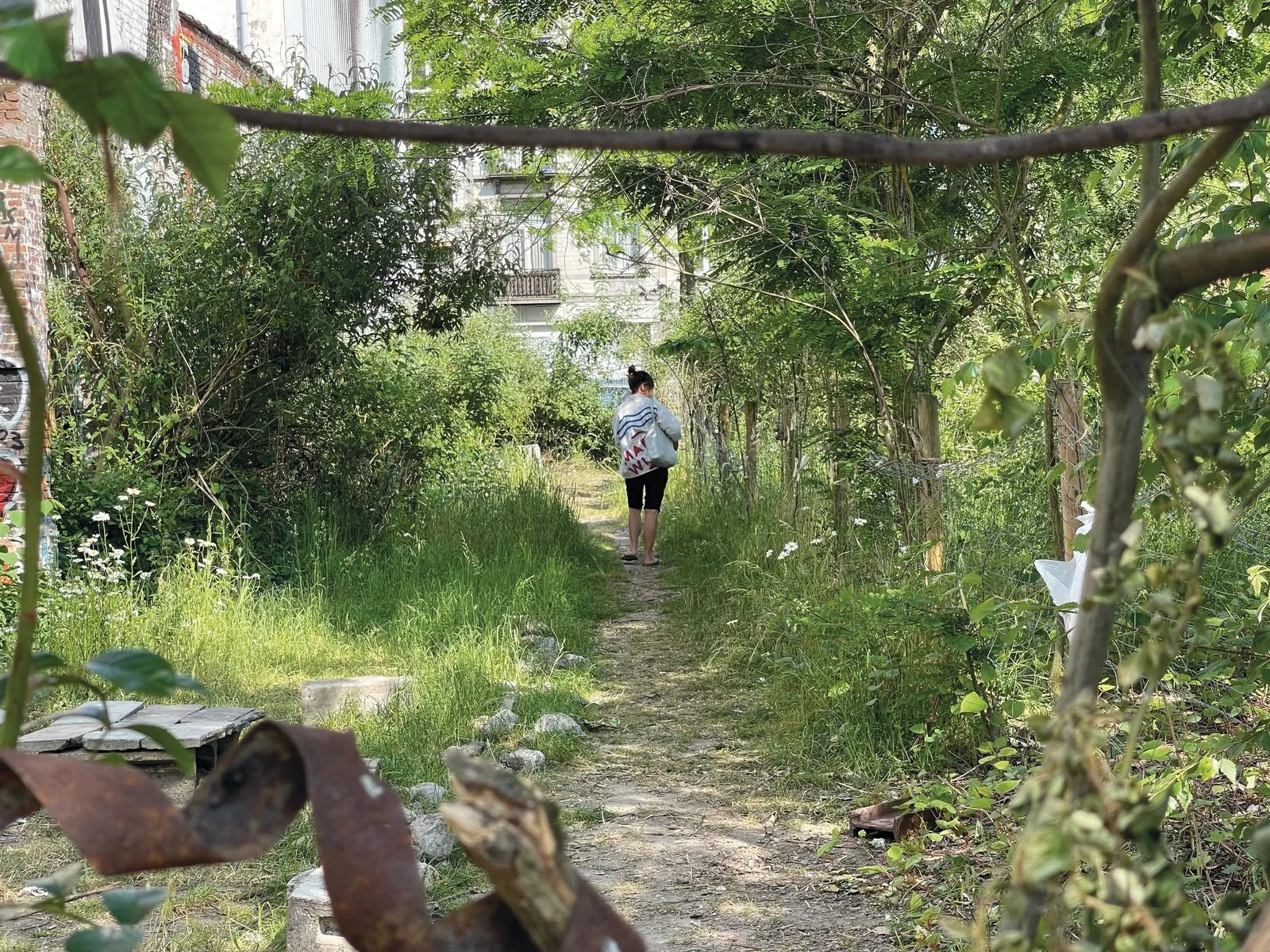 Iemand loopt met een tas over een overwoekerd pad op een Brussels braakliggend terrein.