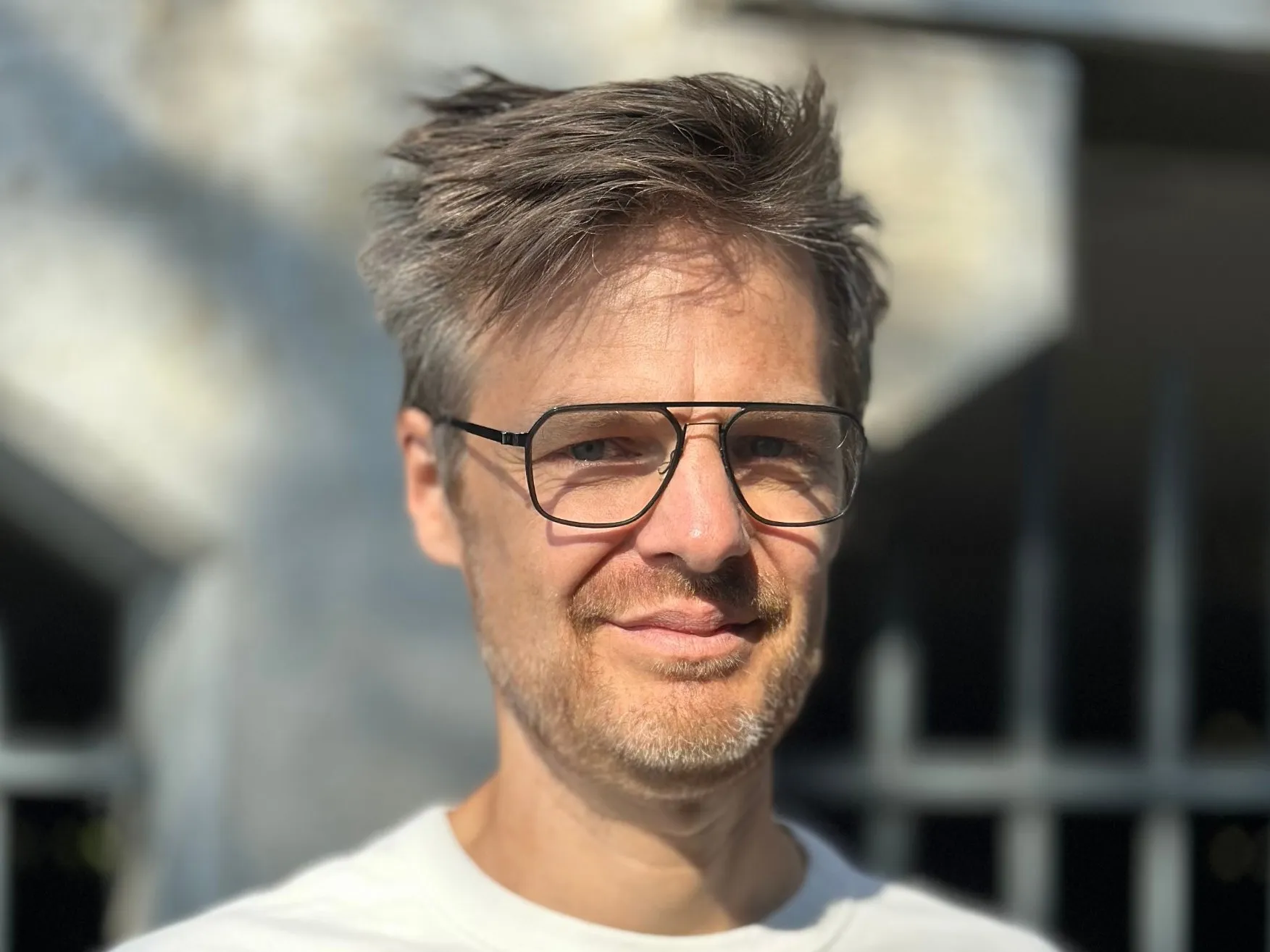 Thomas Laureyssens in een wit T-shirt en met bril, glimlachend voor een zonbeschenen betonnen muur.