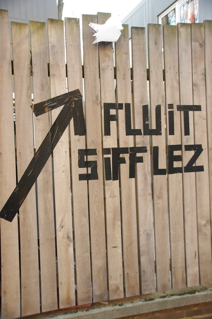 Op een houten schutting staat met zwarte tape een pijl en de woorden FLUIT SIFFLEZ.