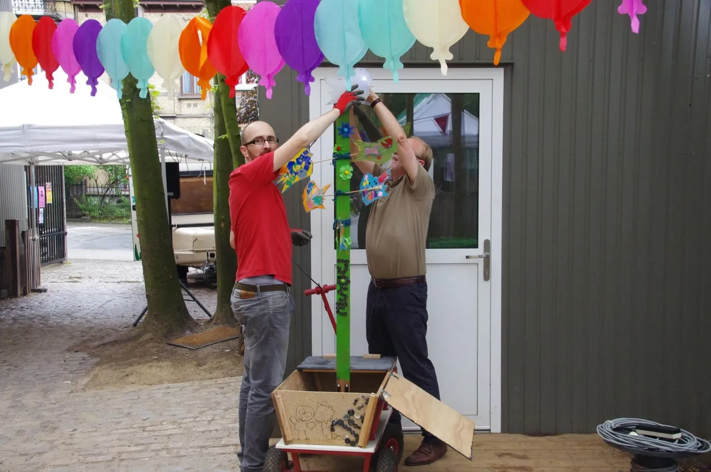 Twee mannen monteren een groene paal met een mus erop, op een houten karretje in een binnenkoer.