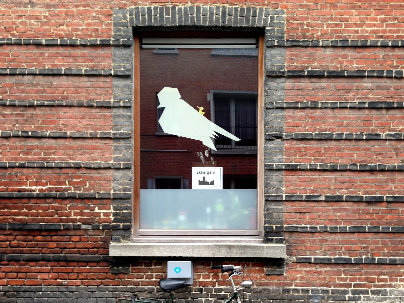 Een witte mus-uitsnede in een raam, met een bordje Ekkergem eronder.